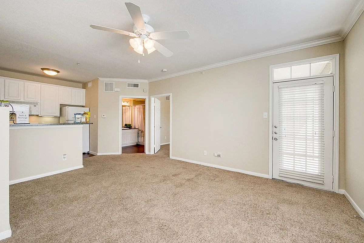 Bright and airy living room with plush carpet, ceiling fan, and an open kitchen with white cabinetry at Stonebriar of Frisco Apartments.