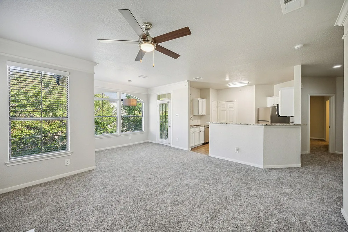 Bright and airy living space with plush carpeting, ceiling fan, large windows, and an open kitchen with modern cabinetry at Stonebriar of Frisco Apartments.