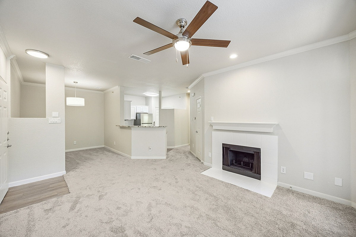 Chic living room at Stonebriar of Frisco Apartments with plush carpeting, elegant ceiling fan, and cozy fireplace, adjacent to a modern kitchen with granite countertops.