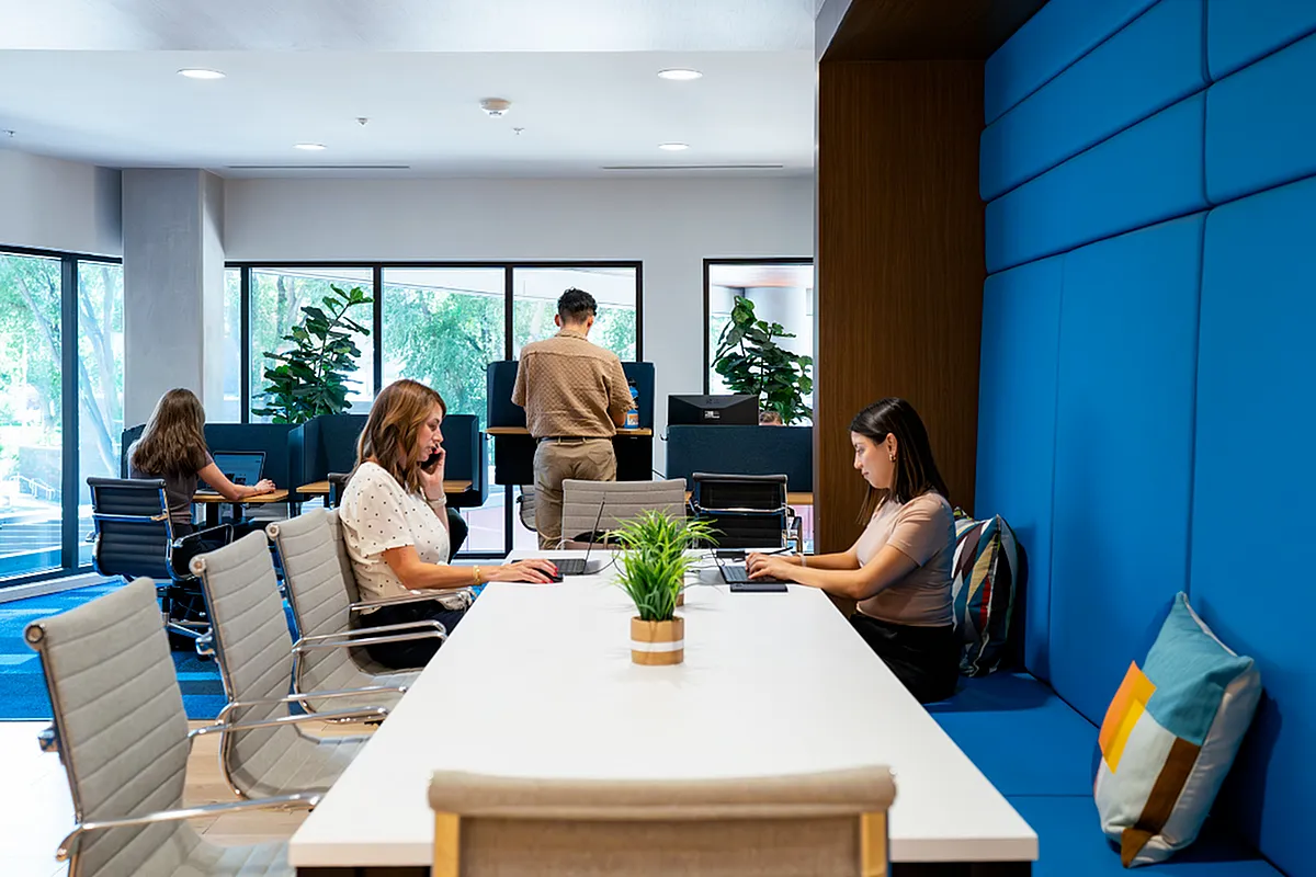 Modern coworking space with bright interiors, featuring professionals engaged in work at a long white table, surrounded by large windows and vibrant blue accents  at Apt CoWork at Sugarmont Apartments in SLC