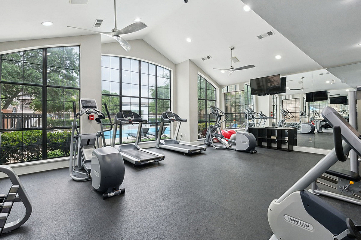 Spacious gym with large windows, featuring treadmills and elliptical machines, offering a vibrant workout environment with a pool view at Toscana at Valley Ridge.