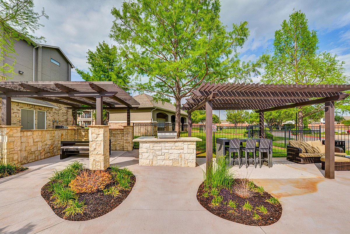 Charming outdoor patio at Toscana at Valley Ridge Apartments with stone BBQ area, stylish pergola, and lush greenery, perfect for entertaining in a serene setting.