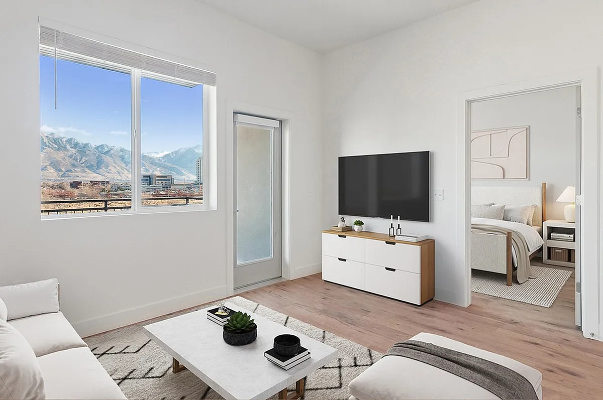 Modern living room at Current Apartments with elegant decor, large window showcasing stunning mountain views, flat-screen TV, and cozy seating area leading into a stylish bedroom.