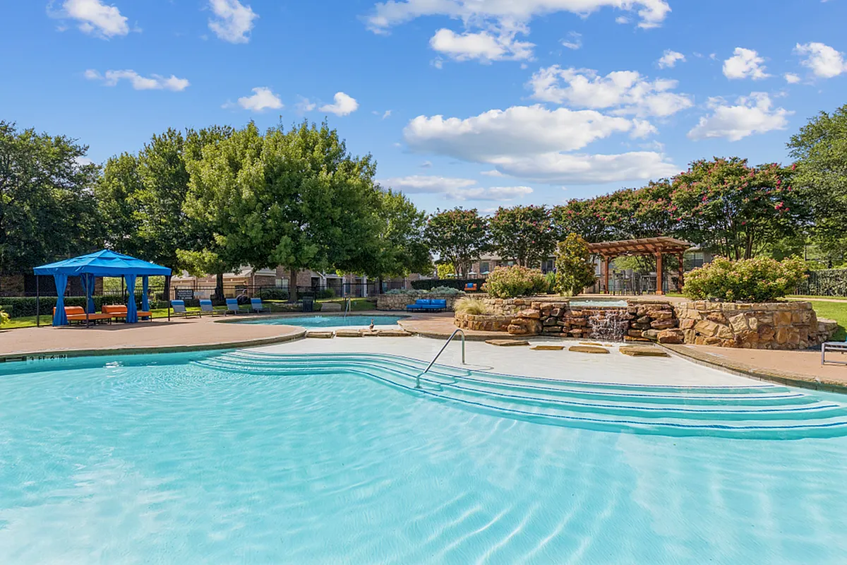 Inviting outdoor swimming pool with cascading waterfall and cozy cabanas, set against lush trees and a vibrant sky at Enclave on Golden Triangle in Forth Worth Texas
