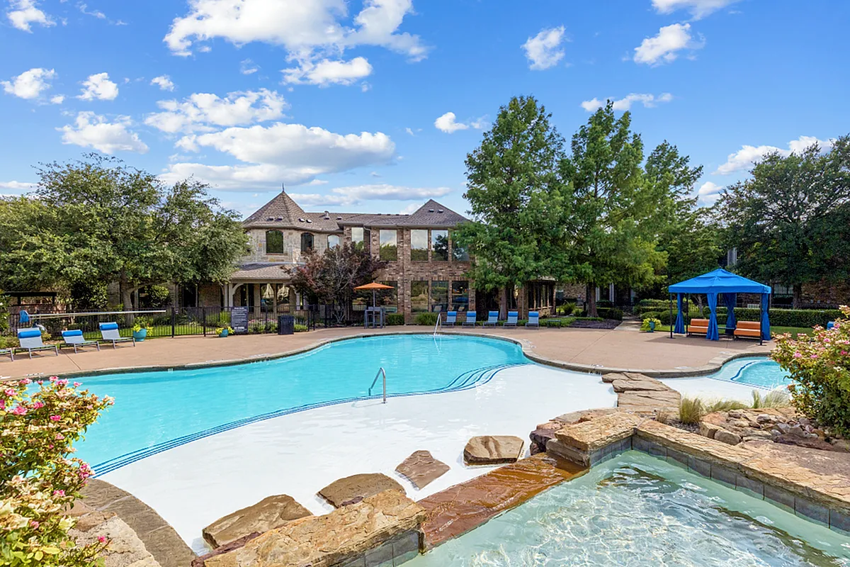 Luxurious apartment complex featuring a sparkling swimming pool, cozy outdoor lounge with blue canopy, and lush landscaping under a clear blue sky at Enclave on Golden Triangle in Forth Worth Texas