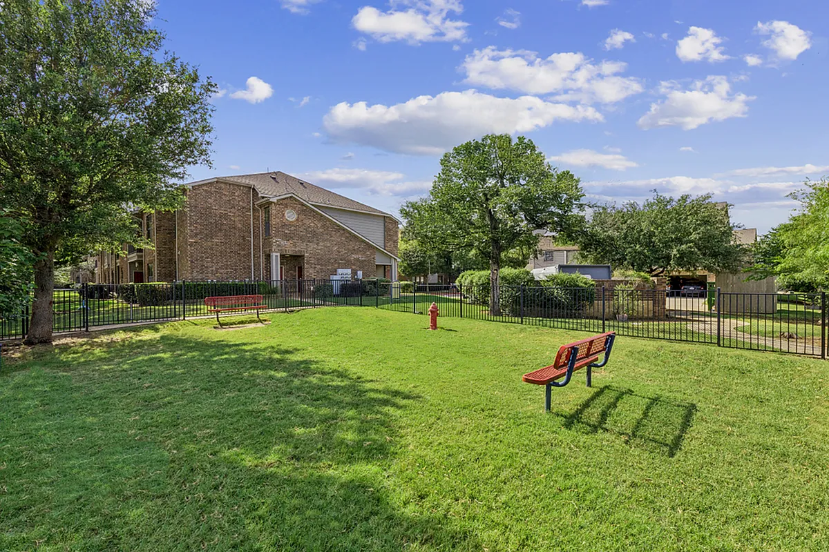 Charming apartment exterior with lush green lawn, benches, and mature trees under a bright blue sky. Serene and inviting apartment complex at Enclave on Golden Triangle in Forth Worth Texas