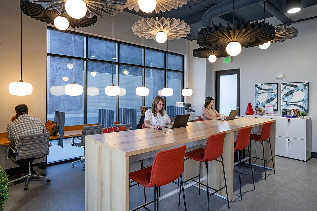 Modern workspace at Park Avenue with vibrant decor, featuring a long wooden communal table, stylish red chairs, and contemporary lighting. Large windows offer ample natural light, creating an inviting and productive environment.