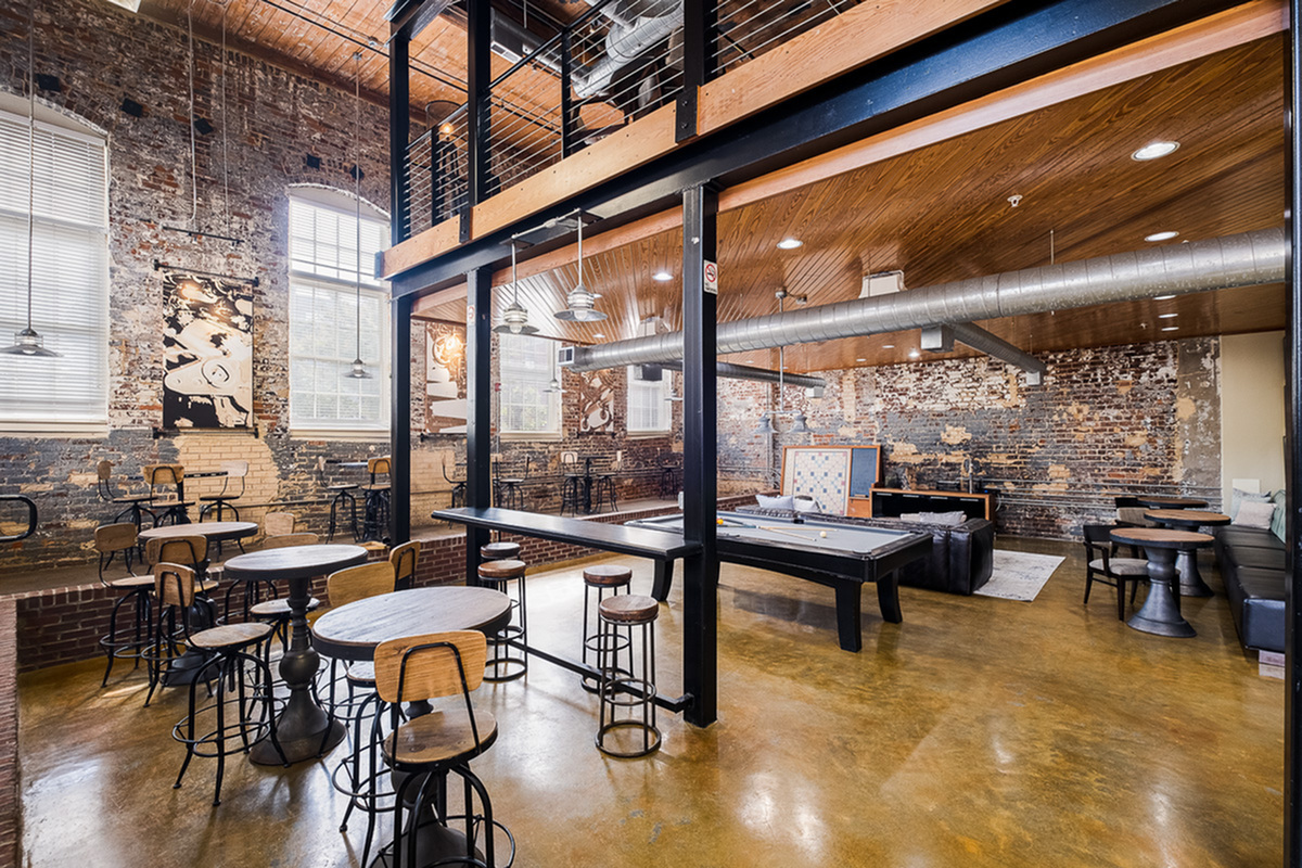 Chic loft-style community area at Alpha Mill Apartments with exposed brick walls, high ceilings, and a blend of modern and industrial design. Features cozy seating, a billiard table, and vibrant natural lighting for an inviting atmosphere.