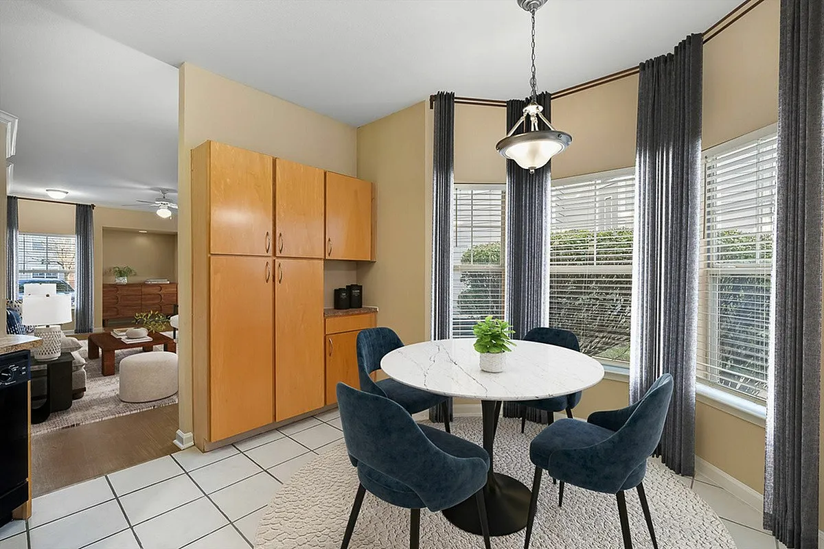 Charming dining area at Cason Estates with a round marble table and elegant blue chairs, surrounded by large windows with lush greenery views, leading to a cozy living room with modern furnishings.
