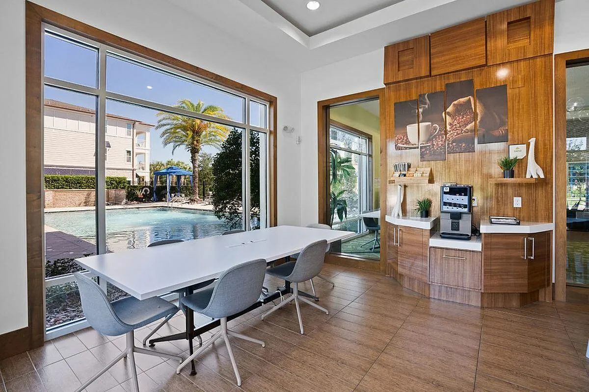 Meeting area at coworking space with sleek wooden decor and a coffee bar overlooking a serene pool with palm trees at Apt CoWork at The Marq Highland Park in Westchase FL