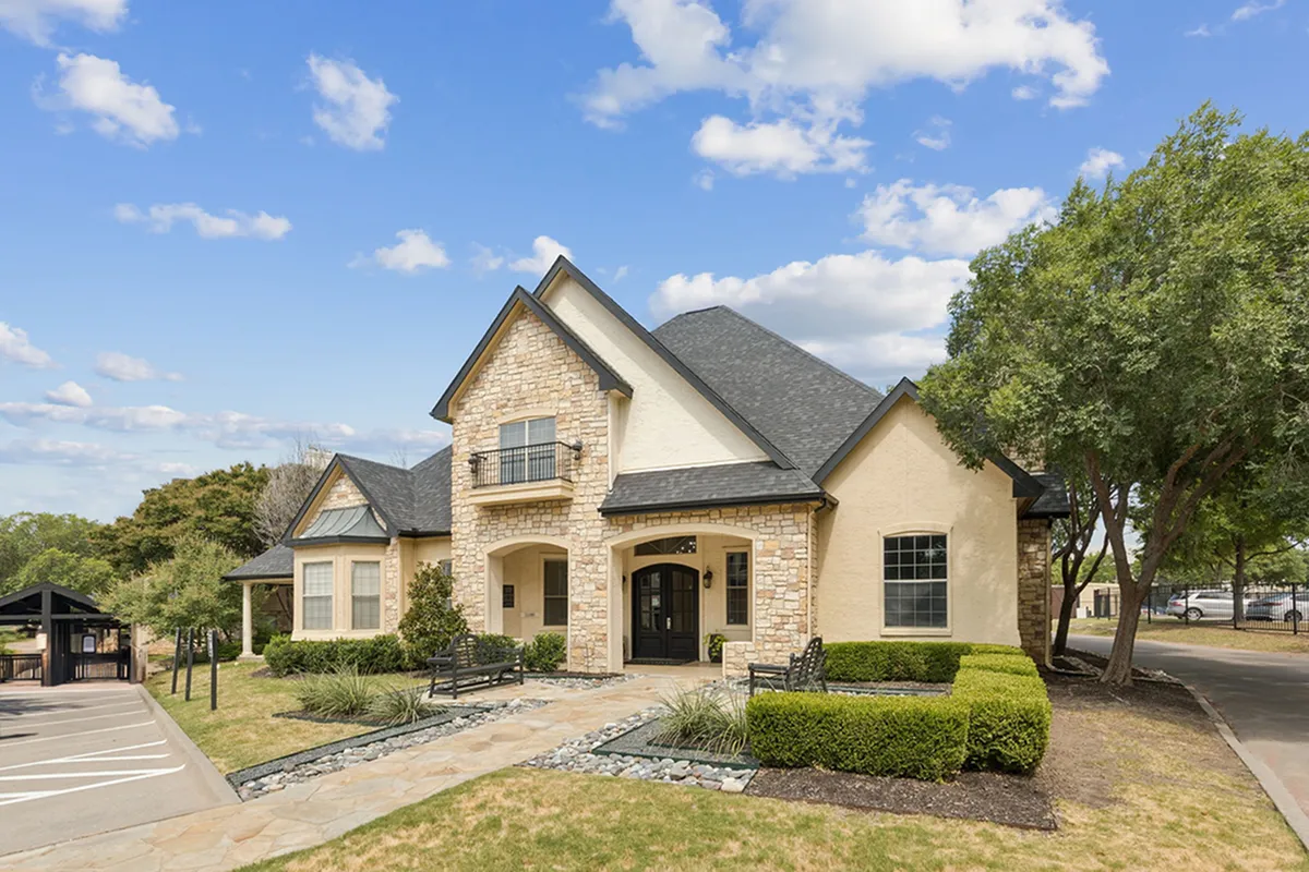 Charming stone apartments with elegant arched entrance, surrounded by lush greenery under a bright blue sky at Stonebriar at Frisco Apartments in Frisco Texas
