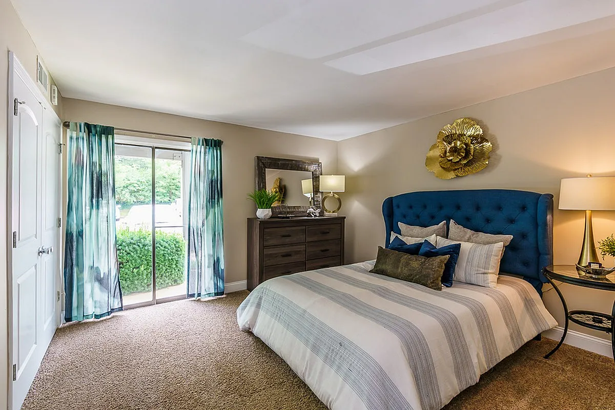 Inviting bedroom at The Creeks on Kirklevington Apartments with elegant decor, featuring a plush blue headboard, stylish striped bedding, and abundant natural light from a large window.