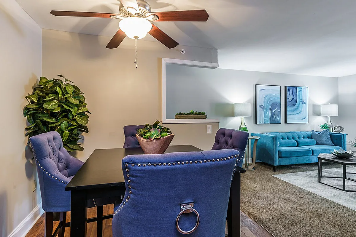 Inviting open-plan living and dining area with blue velvet chairs, a stylish table, and an elegant azure sofa accentuated by modern art and abundant natural light at The Creeks on Kirklevington Apartments.