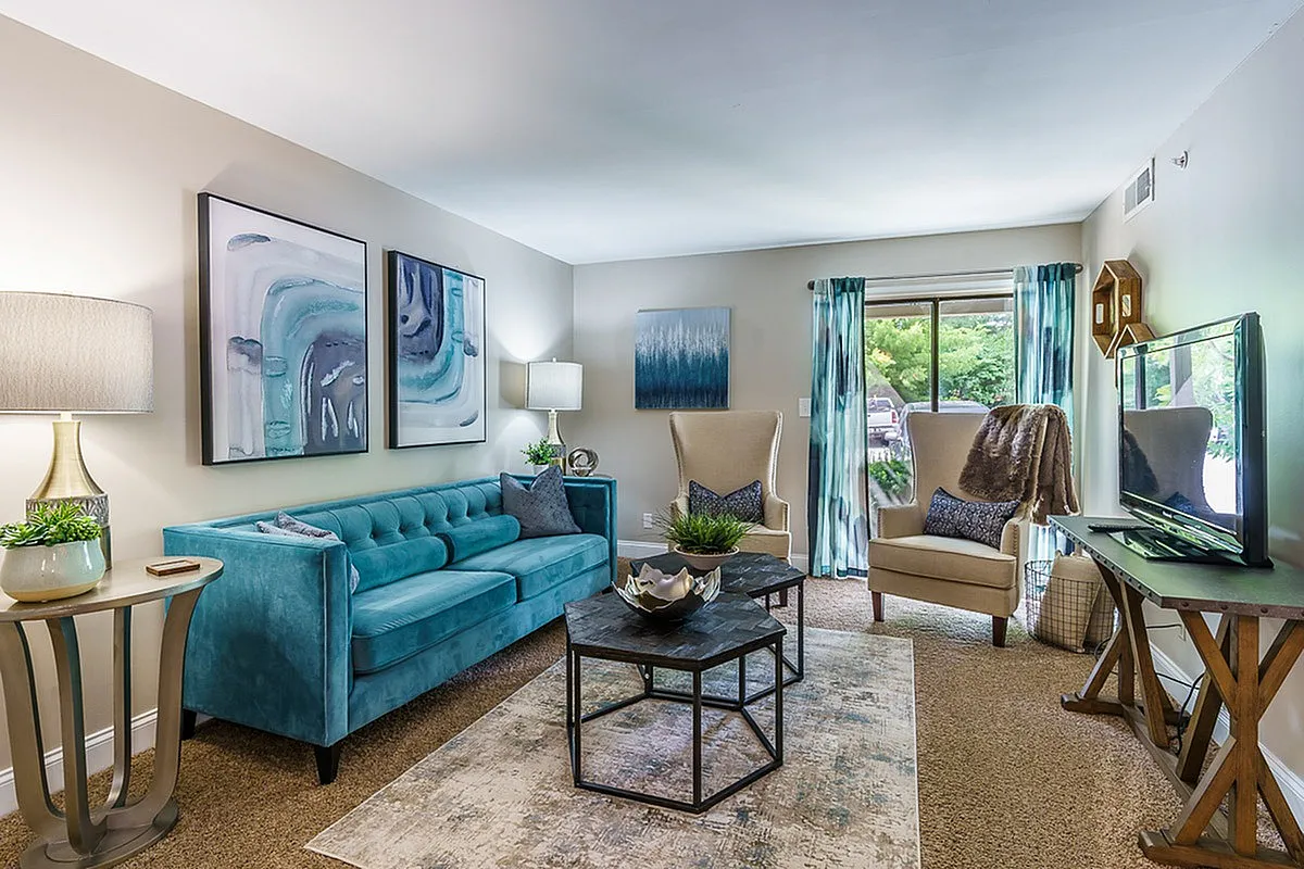 Chic living room at The Creeks on Kirklevington Apartments with elegant teal sofa, stylish armchairs, artistic wall decor, and a cozy atmosphere.