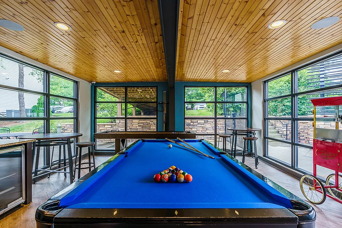 Chic recreation room at The Creeks on Kirklevington Apartments with a vibrant blue pool table, modern wooden ceiling, and large windows offering lush garden views.