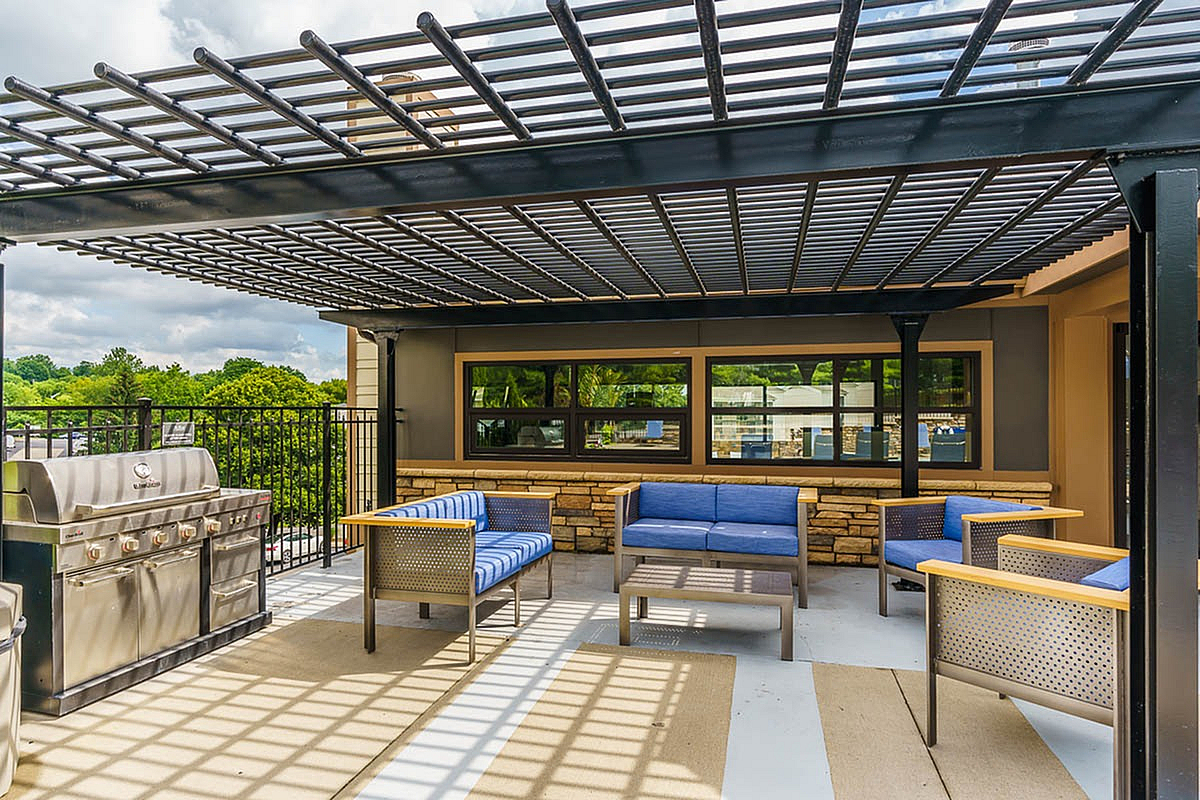 Elegant patio at The Creeks on Kirklevington Apartments featuring modern seating area with blue cushions and a state-of-the-art grill, framed by lush greenery and stylish overhead pergola.
