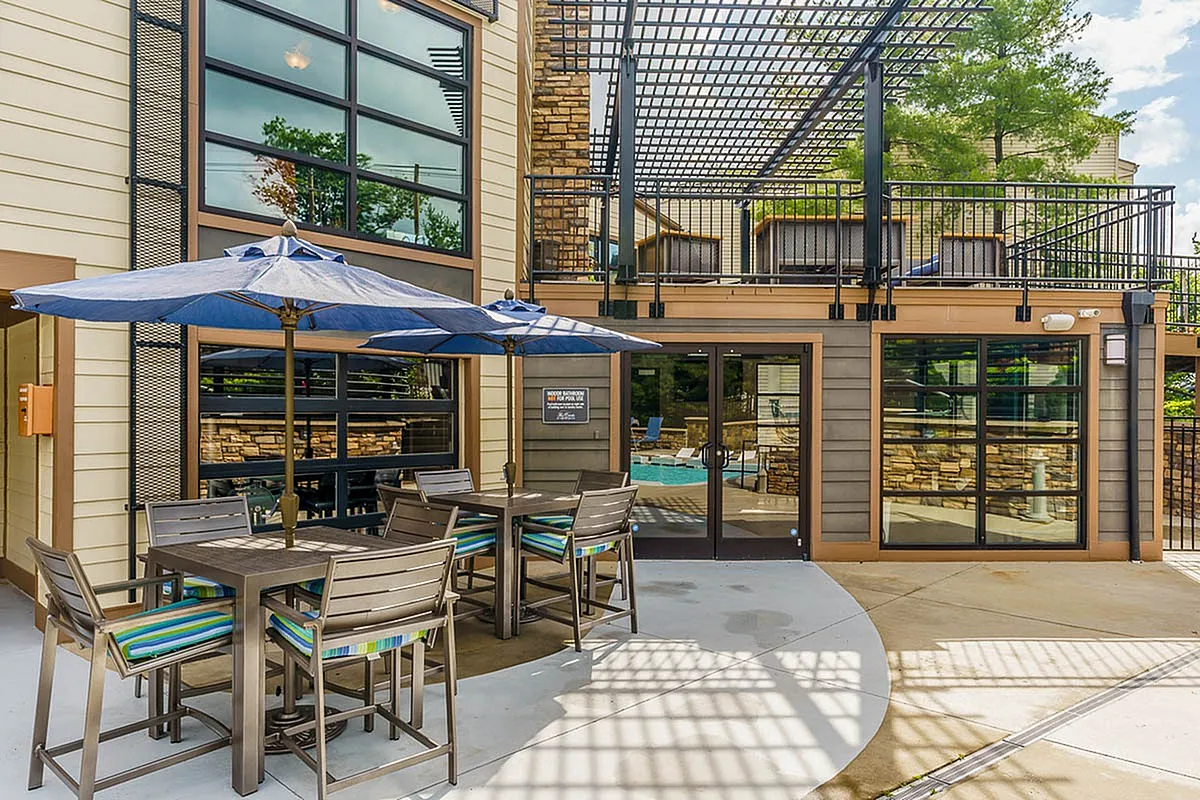Charming outdoor patio at The Creeks on Kirklevington Apartments with modern seating, vibrant blue umbrellas, and a view of inviting pool access beneath sleek architectural design.