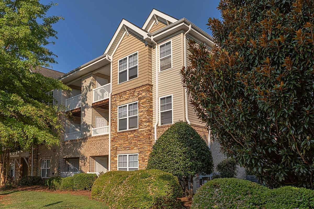 Charming multi-story apartment building, The Stratford at Hillcrest Towne Center Apartments, with brick and stone facade, surrounded by lush greenery and shaded balconies.