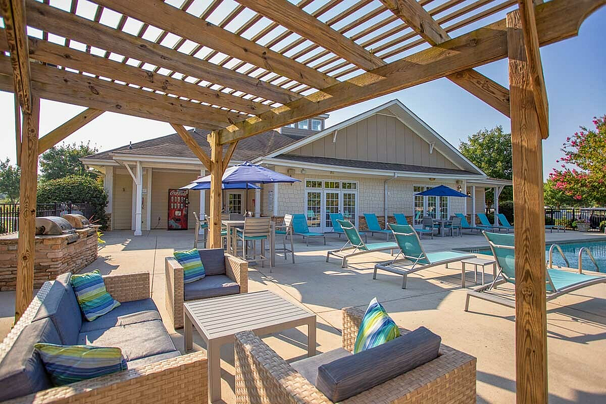 Charming outdoor patio at The Stratford at Hillcrest Towne Center Apartments with wicker furniture, vibrant blue loungers, and a sunlit pergola beside a refreshing pool.