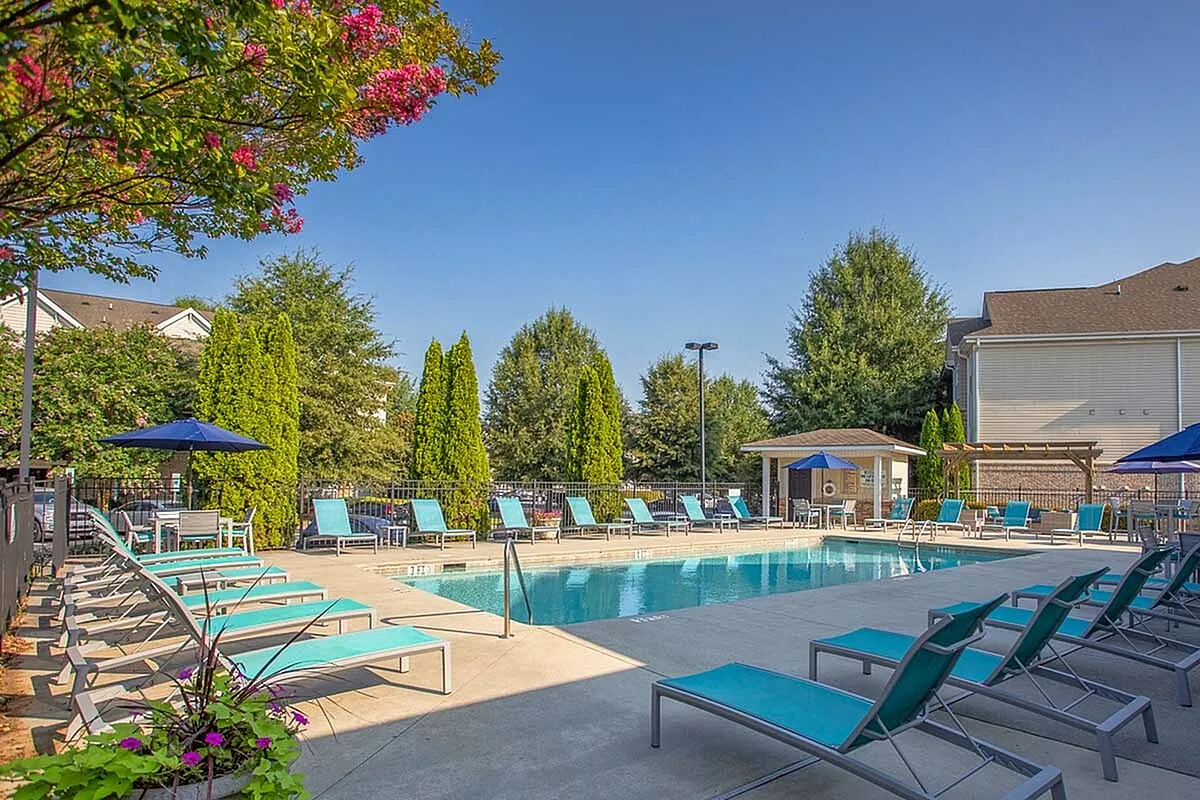 Elegant swimming pool area at The Stratford at Hillcrest Towne Center Apartments with turquoise loungers, vibrant greenery, and a cozy seating pavilion under a clear blue sky.