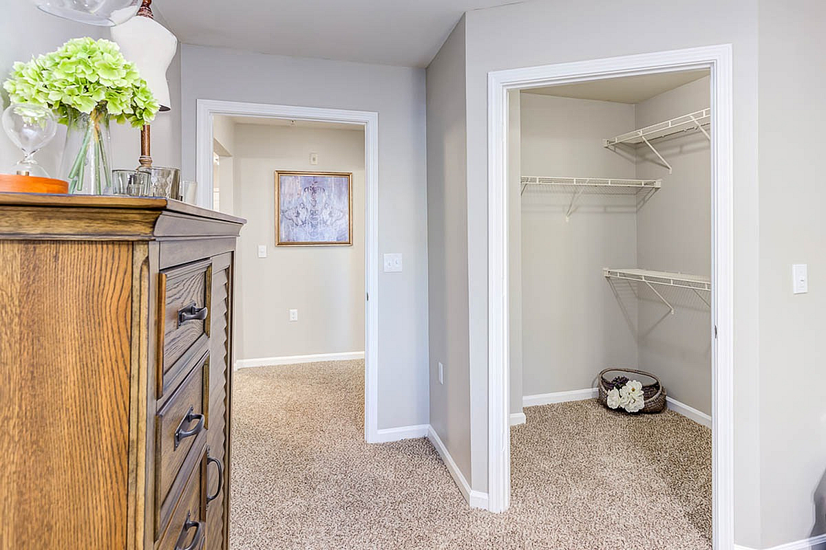 Inviting apartment interior featuring a cozy hallway, elegant wooden dresser with decorative accents, and a spacious open closet with plush carpet flooring, perfect for stylish living at The Stratford at Hillcrest Towne Center Apartments.