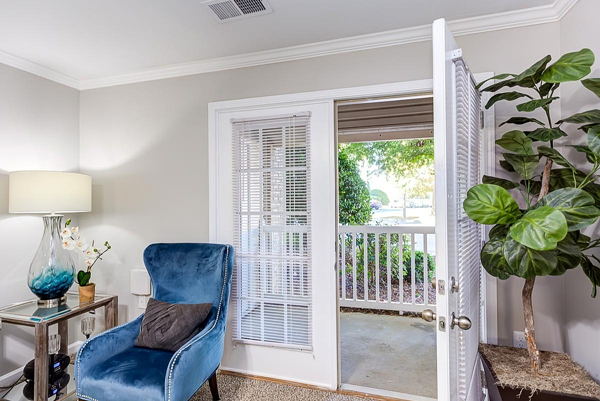 Inviting living space at The Stratford at Hillcrest Towne Center Apartments with elegant blue armchair, stylish lamp, and private patio access through glass doors, complemented by greenery for a serene ambiance.
