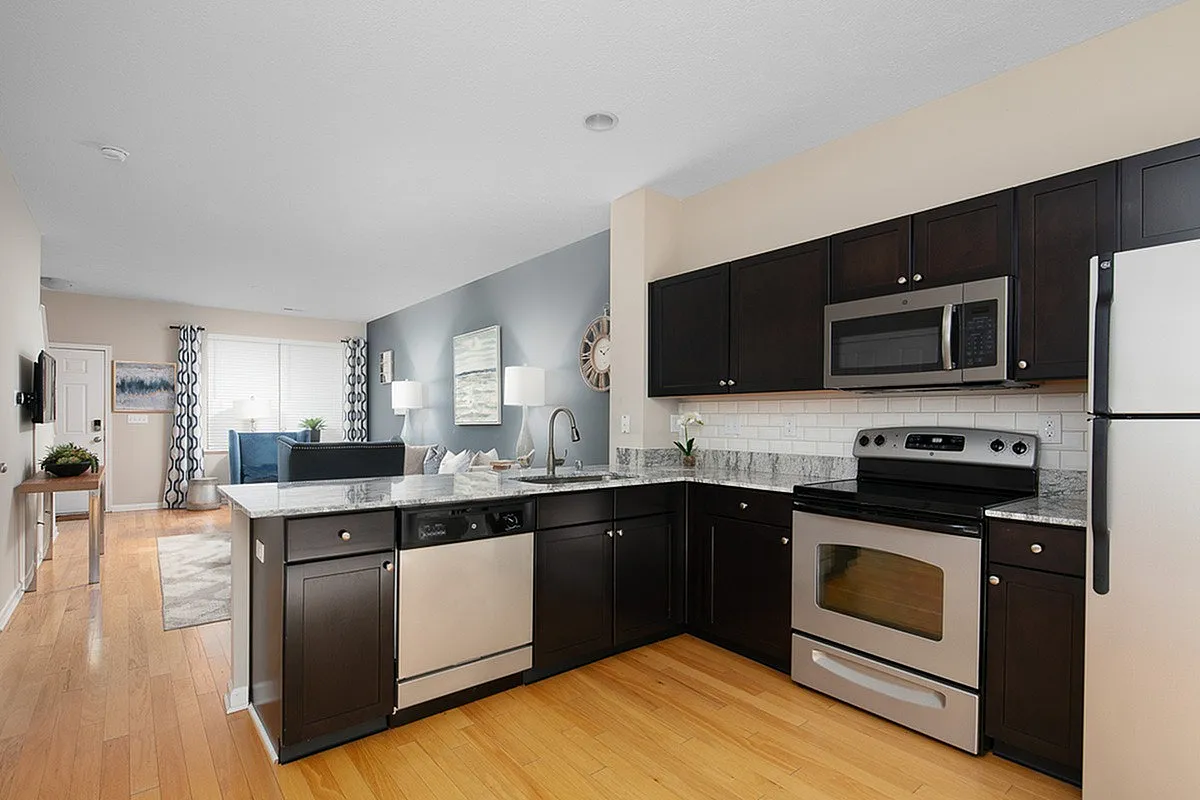 Contemporary open-plan kitchen and living space at The Mill at Georgetown Apartments with stylish dark cabinetry, stainless steel appliances, and hardwood flooring.