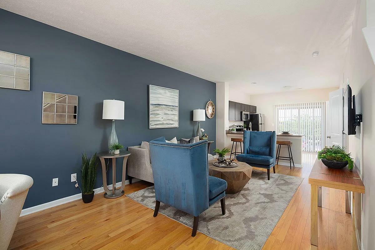 Modern living room at The Mill at Georgetown Apartments with plush blue chairs, warm wooden flooring, and stylish decor, leading into a sleek open kitchen with natural light.