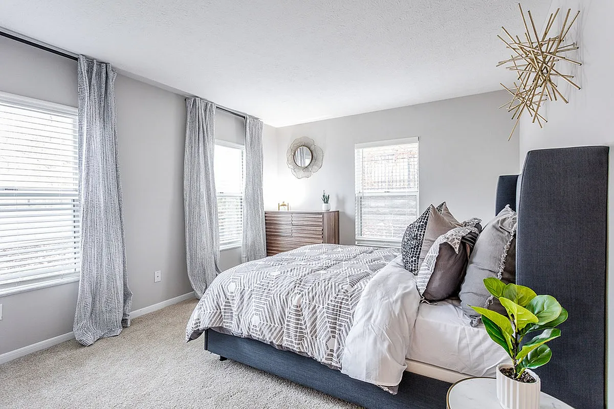 Bright and modern bedroom at The Retreat at Stillmeadow Apartments with elegant decor, featuring a plush gray bed, stylish pillows, and a wooden dresser. Natural light flows through large windows with chic gray curtains, enhancing the room's serene ambiance.