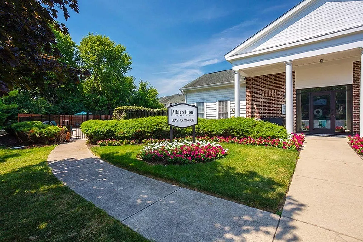 Charming entrance to Alkire Glen Apartments leasing office surrounded by vibrant flower beds and lush greenery under a clear blue sky.