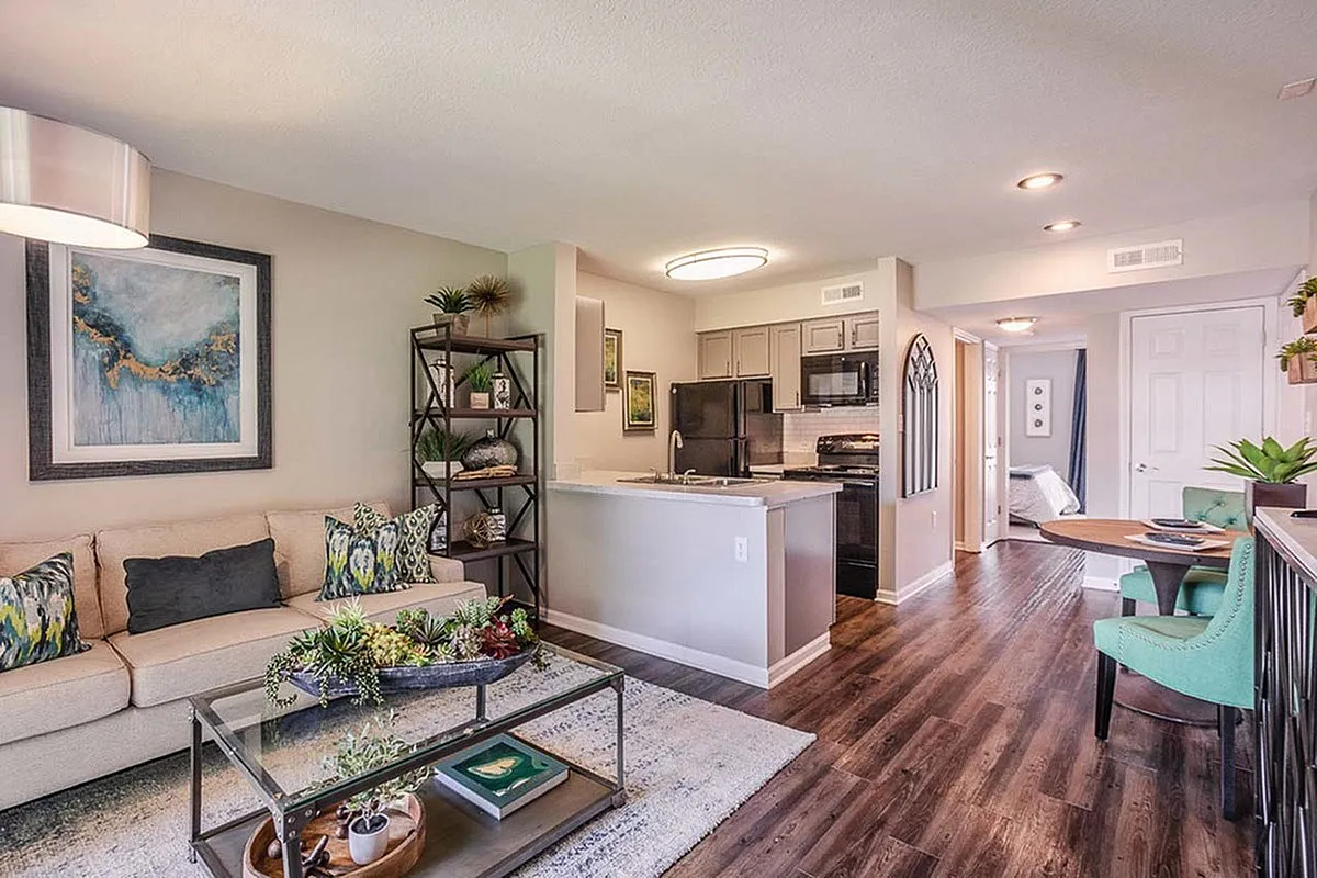 Elegant open-plan living room with stylish decor, featuring a cozy beige sofa, vibrant artwork, and a modern kitchen with sleek appliances at Alkire Glen Apartments.