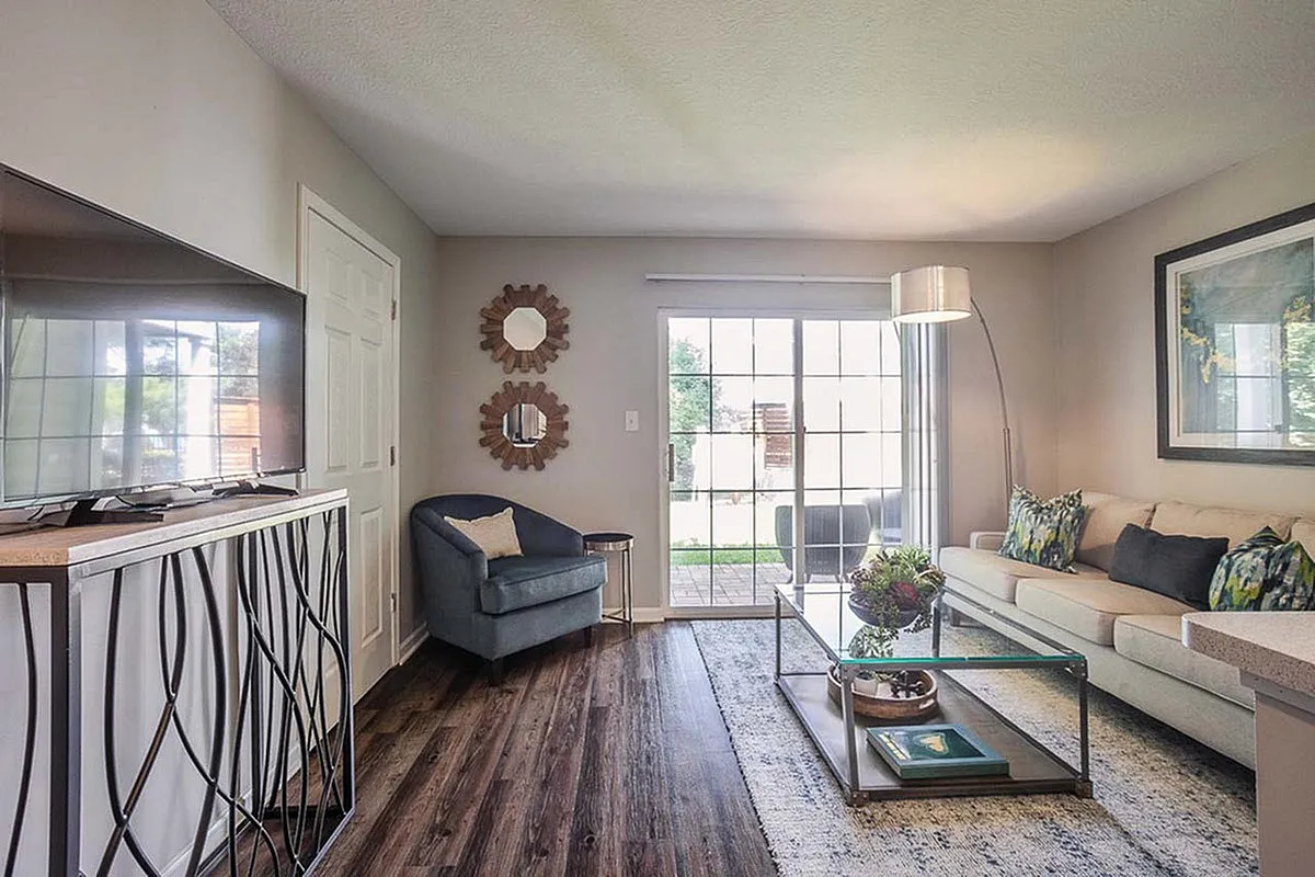 Elegant living room at Alkire Glen Apartments featuring a cozy armchair, stylish mirror decor, and bright glass doors leading to a lush outdoor patio.