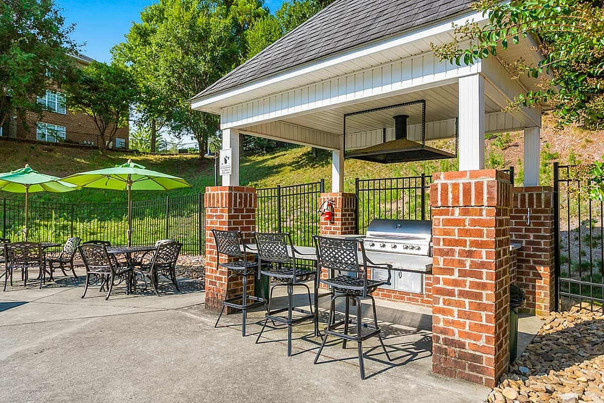 Sunny outdoor barbecue area at Morgan Ridge Apartments with brick grill station, shaded patio seating, and vibrant green umbrellas in a lush setting.