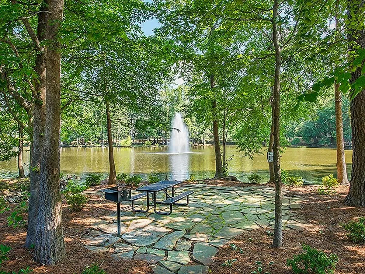 Serene outdoor space at Lake St James Apartments with a stone patio, picnic table, and a view of a tranquil lake with a beautiful fountain, surrounded by lush greenery.