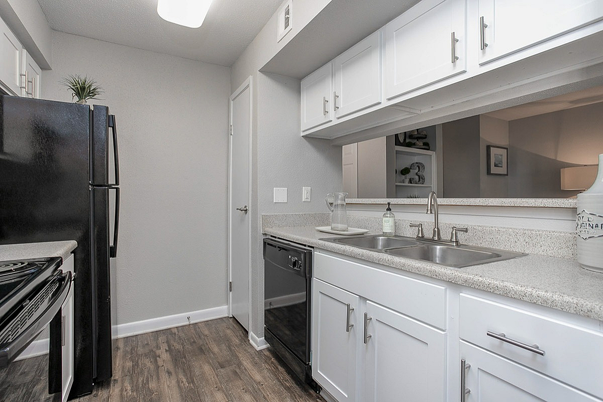 Modern kitchen at Autumn Ridge Apartments with sleek white cabinetry, polished countertops, and stainless steel sink, complemented by elegant dark appliances and chic wood flooring.