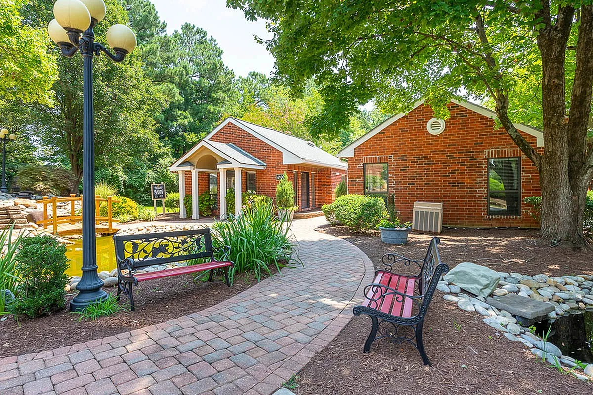 Charming brick clubhouse at Autumn Ridge Apartments surrounded by lush greenery and stone pathways, featuring picturesque benches by a serene pond.