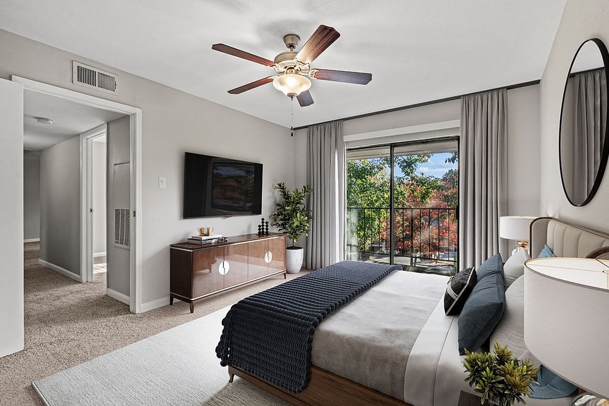 Elegant bedroom with modern decor, featuring a plush bed, flat-screen TV, ceiling fan, and large sliding glass doors leading to a beautiful balcony view at Autumn Ridge Apartments