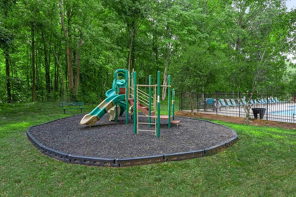 Charming outdoor playground area with vibrant green slide, surrounded by lush trees and next to a refreshing swimming pool, perfect for family enjoyment at Park at Oak Ridge Apartments.