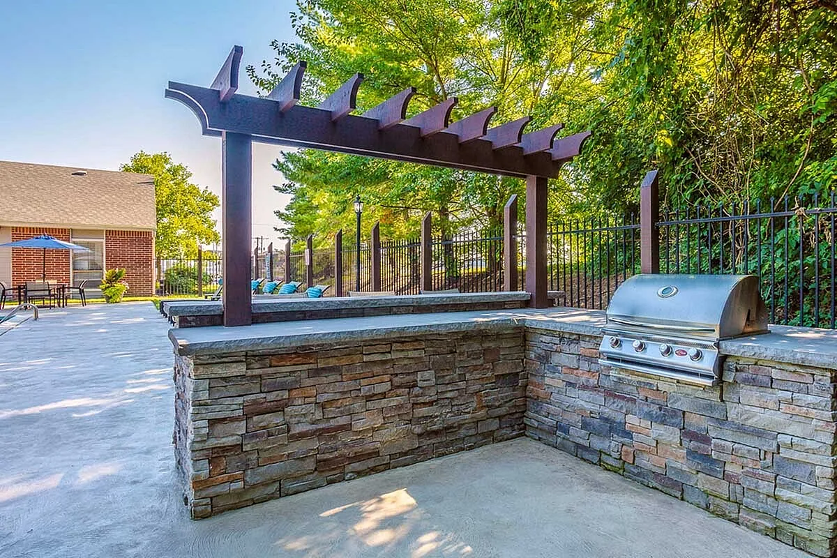 Charming outdoor grilling area at Timber Hollow Apartments with a stone island and modern pergola, surrounded by lush greenery.
