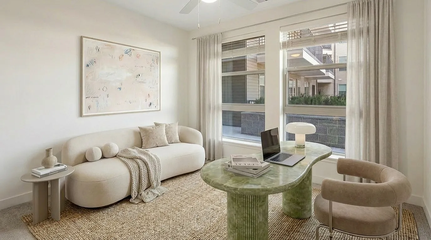 Chic and cozy living room featuring a plush beige sofa, elegant abstract art, and a sleek modern desk with a laptop, bathed in natural light from large windows at The Richmond Apartments in SLC