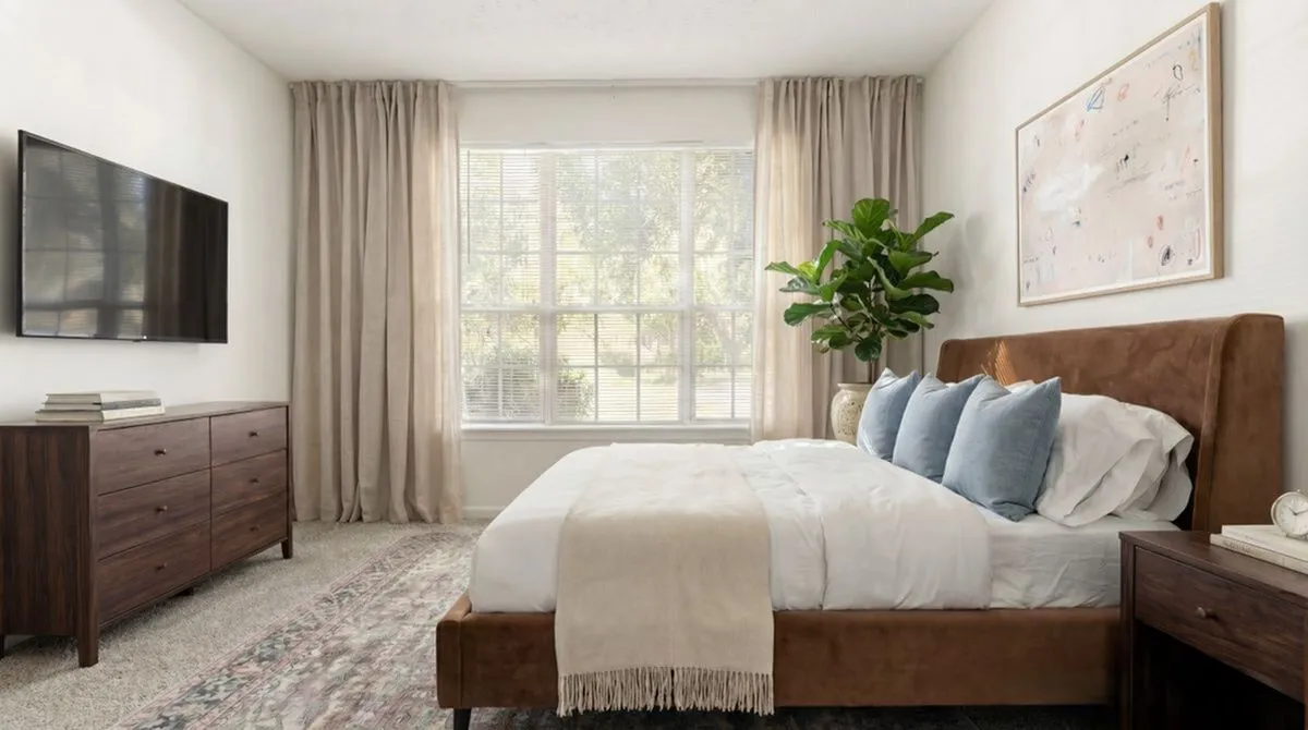 Bright and inviting bedroom with a large window, cozy bed, stylish dresser, and lush plant, perfect for relaxation at Lake St James Apartments in Conyers GA