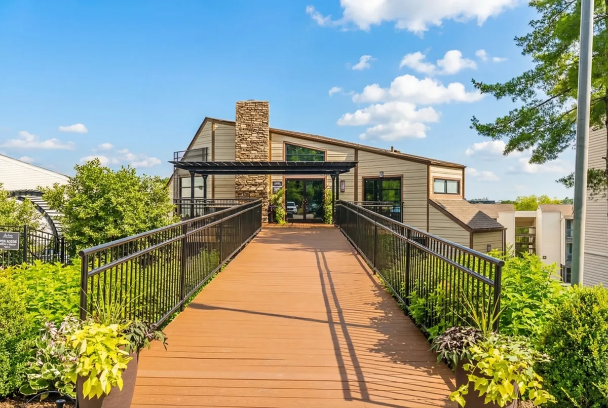 Inviting apartment entrance with a modern walkway surrounded by lush greenery, leading to a stylish building under a bright blue sky.