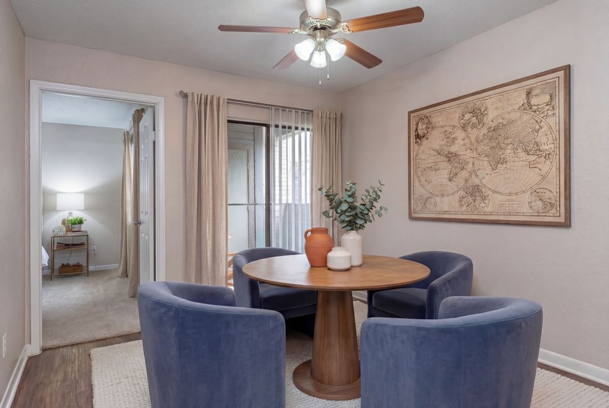 Cozy dining area with a round wooden table, surrounded by elegant blue chairs and illuminated by a stylish ceiling fan. Décor includes a vintage world map and modern pottery accents, creating a warm and inviting atmosphere at Steepleway Downs Apartments in Houston