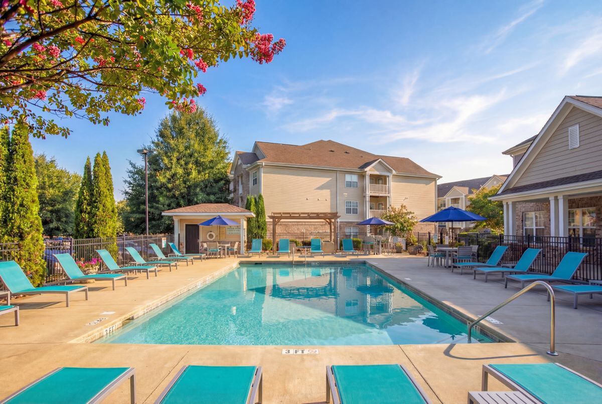 Inviting outdoor pool area with vibrant blue lounge chairs, surrounded by lush greenery and charming apartment buildings at Stratford at Hillcrest Apartments in Winston-Salem.