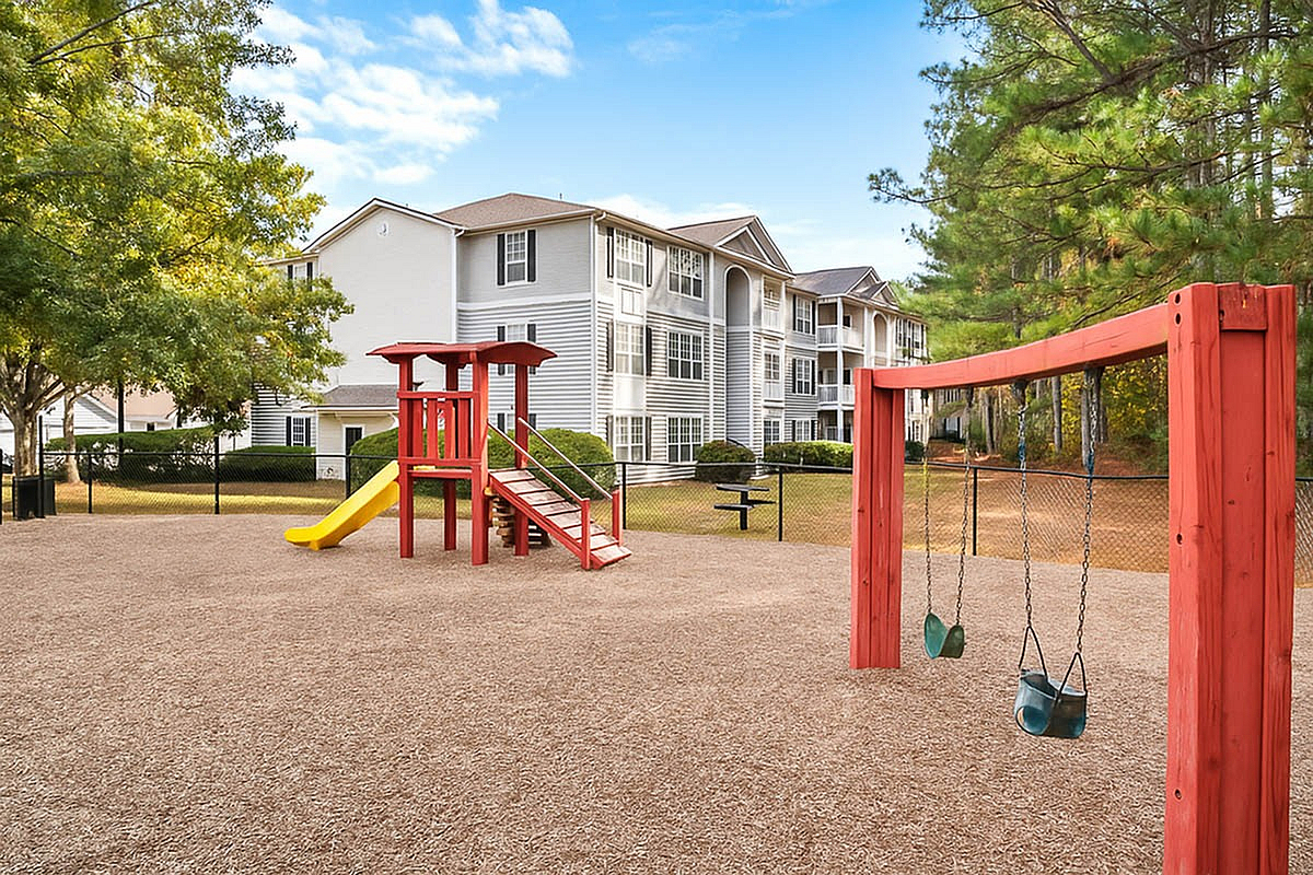 Inviting apartment complex with modern playground featuring a vibrant play structure and swings, surrounded by lush green trees and clear blue skies at Lake St James Apartments in Conyers.
