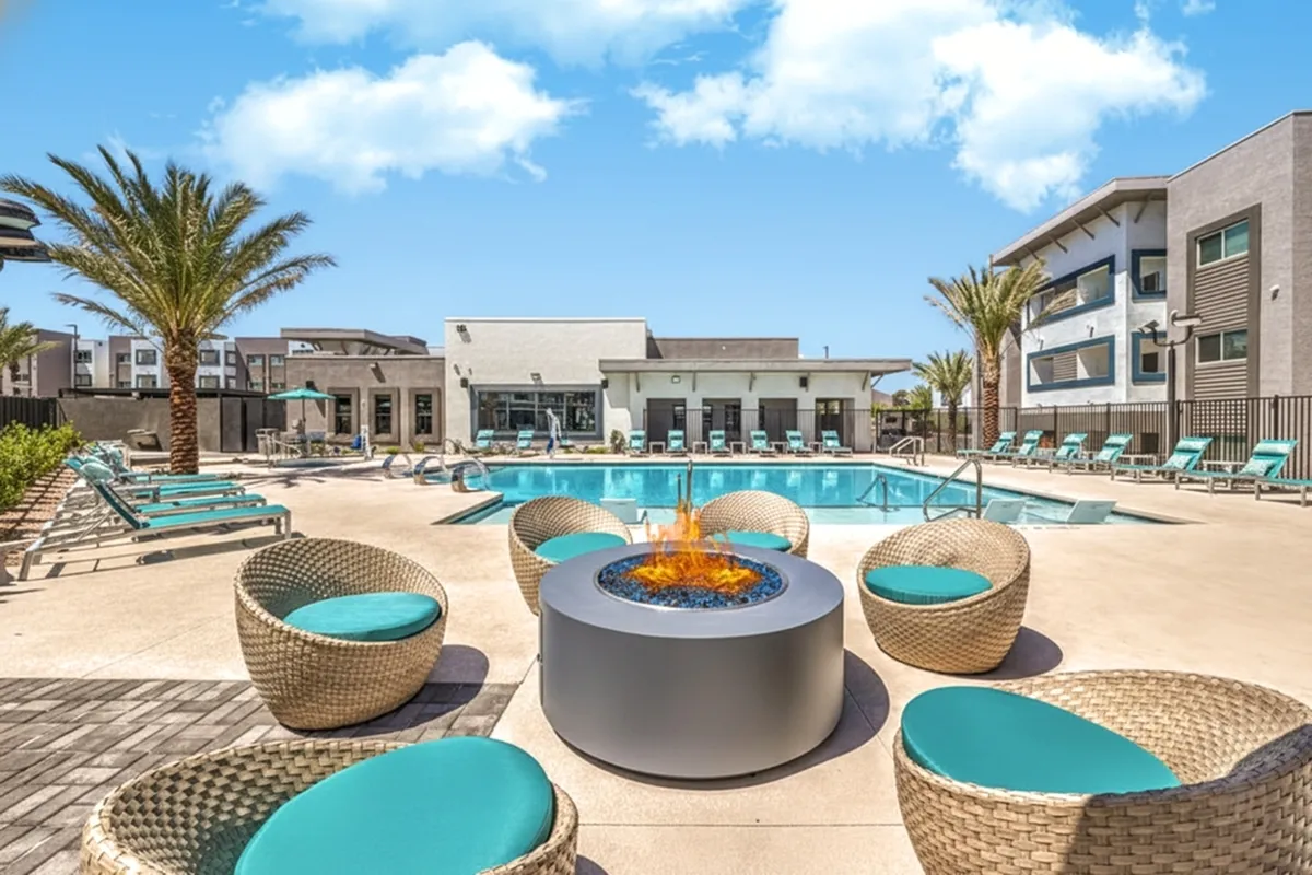 Luxurious apartment complex showcasing a stylish outdoor pool area with cozy wicker seating and a modern fire pit, surrounded by palm trees under a clear blue sky.