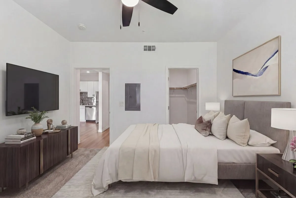 Elegant bedroom showcasing a plush bed with neutral-colored bedding, contemporary decor, and a wall-mounted television, opening into a sleek kitchen area.