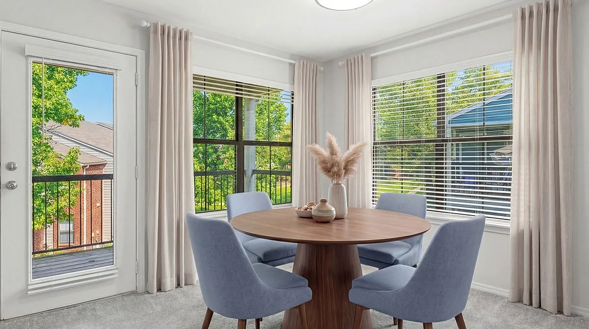 Bright and airy dining nook with modern decor, featuring a round wooden table and plush blue chairs, overlooking a lush green view through large windows at Timber Hollow Apartments