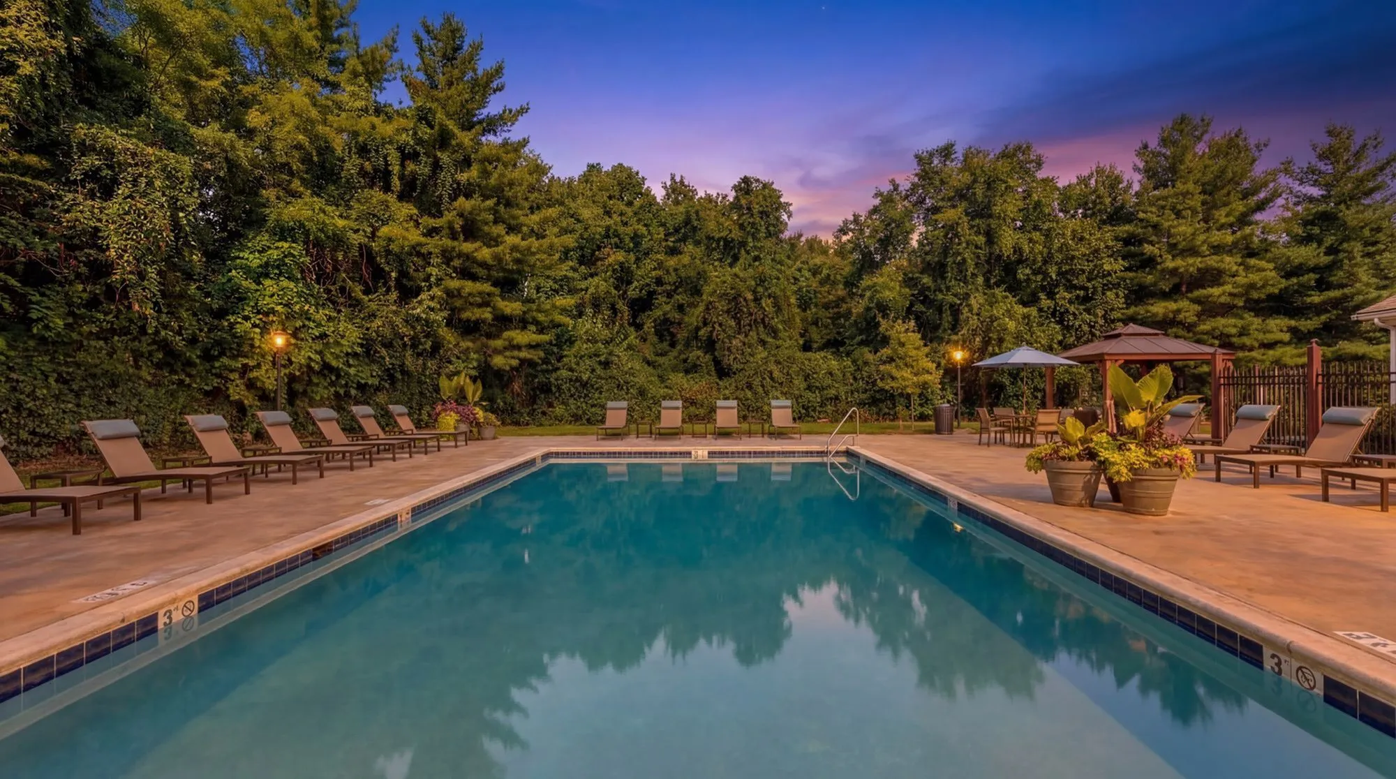 Inviting outdoor pool area surrounded by lush greenery, perfect for relaxation at sunset with comfortable lounge chairs at Timber Hollow Apartments