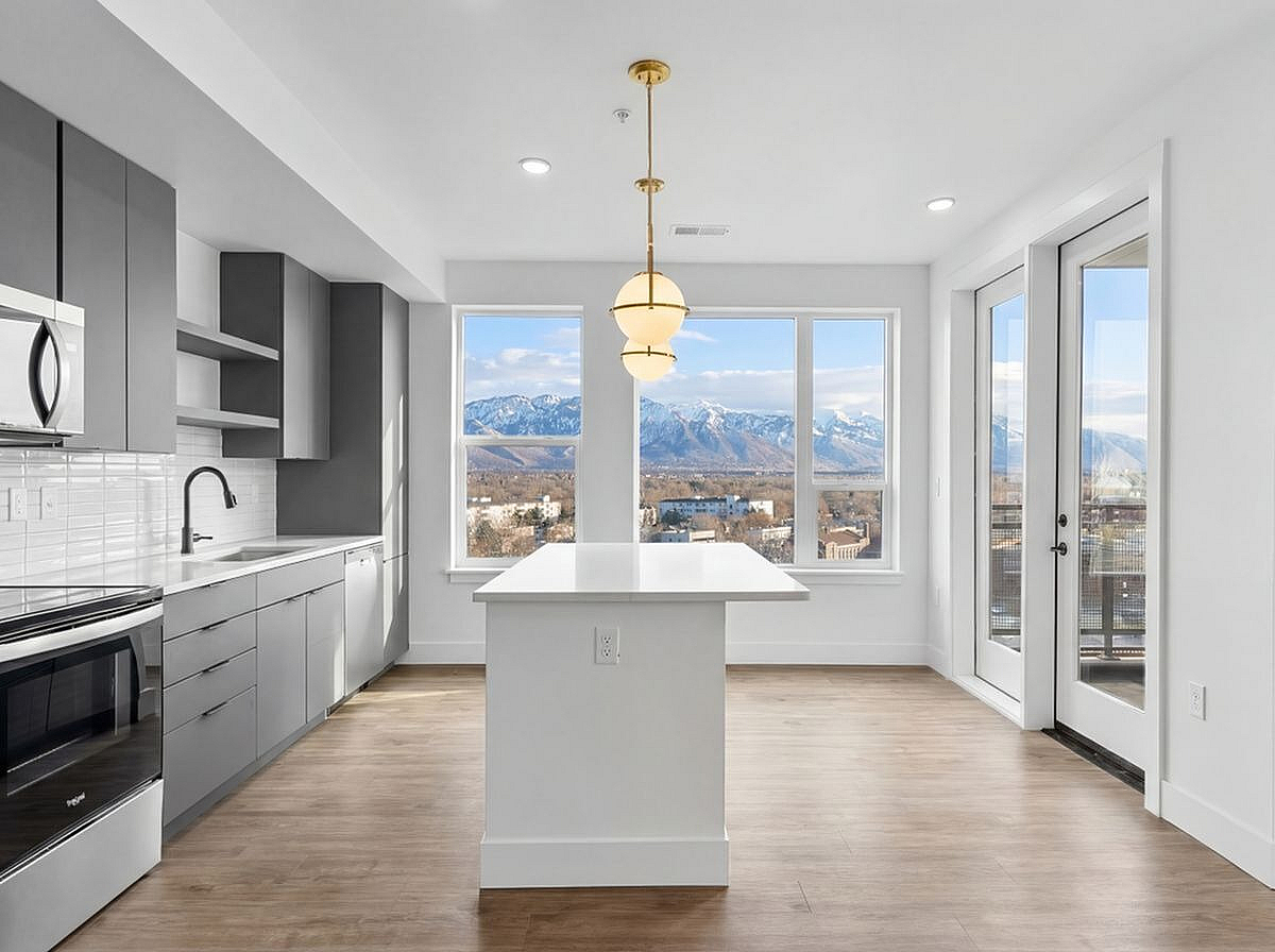 Bright modern kitchen with sleek gray cabinetry, large windows showcasing stunning mountain views, and elegant pendant lighting above a central island kitchen at The Westerly in Millcreek Utah